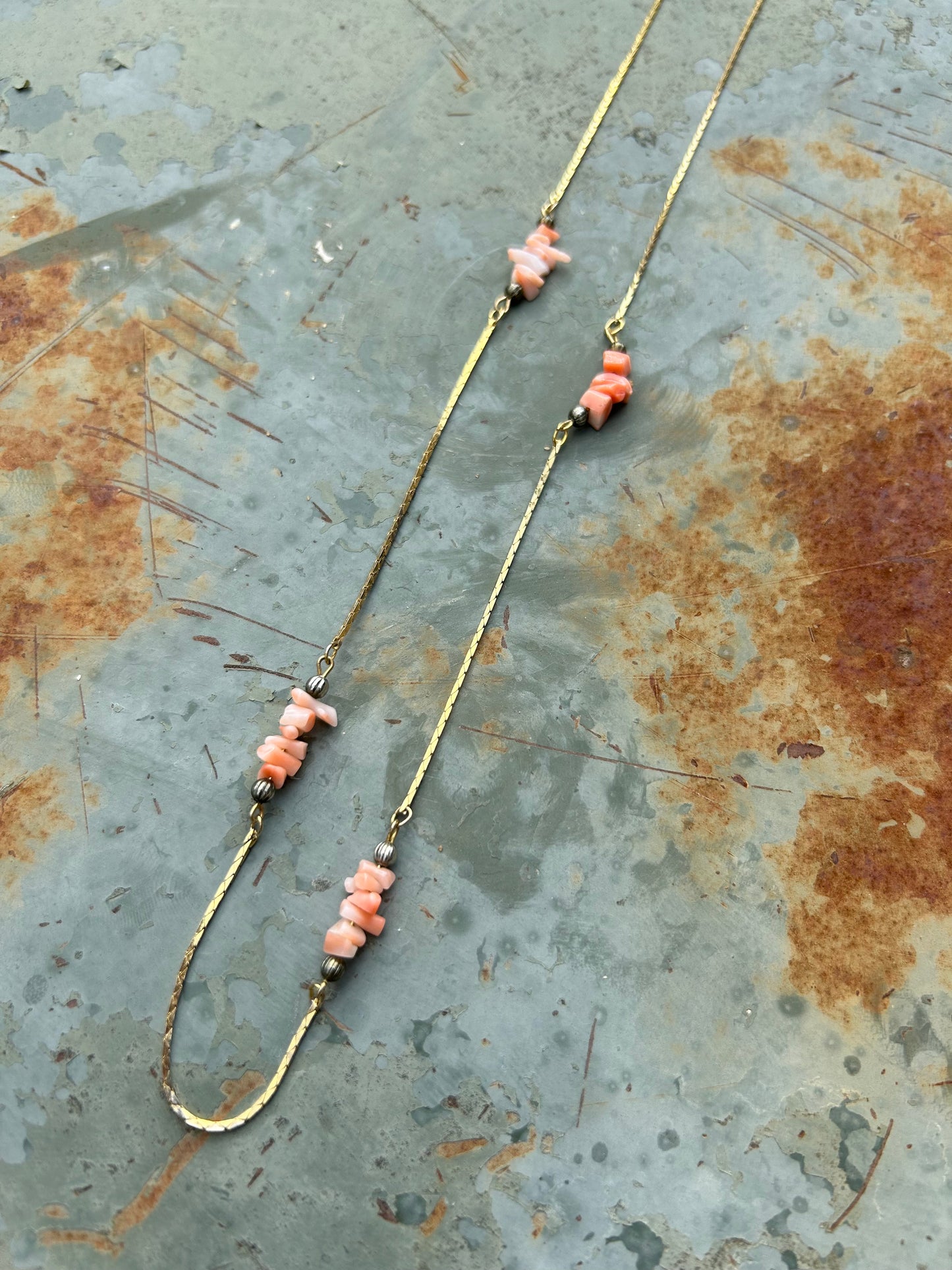 A delicate Canyon Jewelry sterling silver Pink Lady Necklace with pink and coral beads, spaced with small silver beads, laid out on a wooden surface next to decorative pottery and a fringe of a white textile.